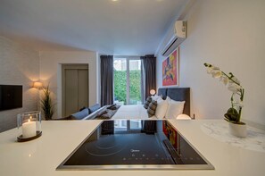 A modern hotel room with a bed, a flat-screen TV, a built-in stove, and a view of greenery outside. - St. Julian's Studio 102 - Near Beach & Paceville (Saint Julian's)