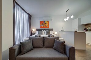 Ein modernes Wohnzimmer mit einer Couch, einem Essbereich und einer Küche im Hintergrund sowie einem großen Fenster mit Vorhängen.