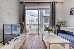 Ein modernes Wohnzimmer mit einem Flachbildfernseher, einem Glastisch, einer Couch und einem Balkon mit Blick auf Gebäude.