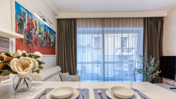 Un coin repas avec une table dressée pour deux personnes, un vase de fleurs, un tableau accroché au mur et une vue sur les bâtiments par la fenêtre.