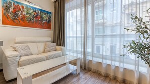 Un salon moderne avec un canapé blanc, une table basse en verre, un tableau au mur et une grande fenêtre ornée de rideaux légers.