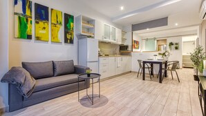 Uma sala de estar moderna com sofá, uma mesa redonda pequena e uma área de jantar com cadeiras. Há também uma cozinha com armários e micro-ondas.