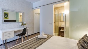 A modern hotel room with a white desk, a black chair, a bed with striped pillows, a glass shower enclosure, and a wooden floor. - St. Julian's Studio 302 - Near Beach & Paceville (Saint Julian's)