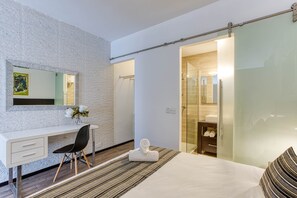 Ein modernes Hotelzimmer mit einem weißen Schreibtisch, einem schwarzen Sessel, einem Bett mit gestreiftem Kopfkissen und einer Duschkabine aus Glas. Der Boden ist aus Holz.
