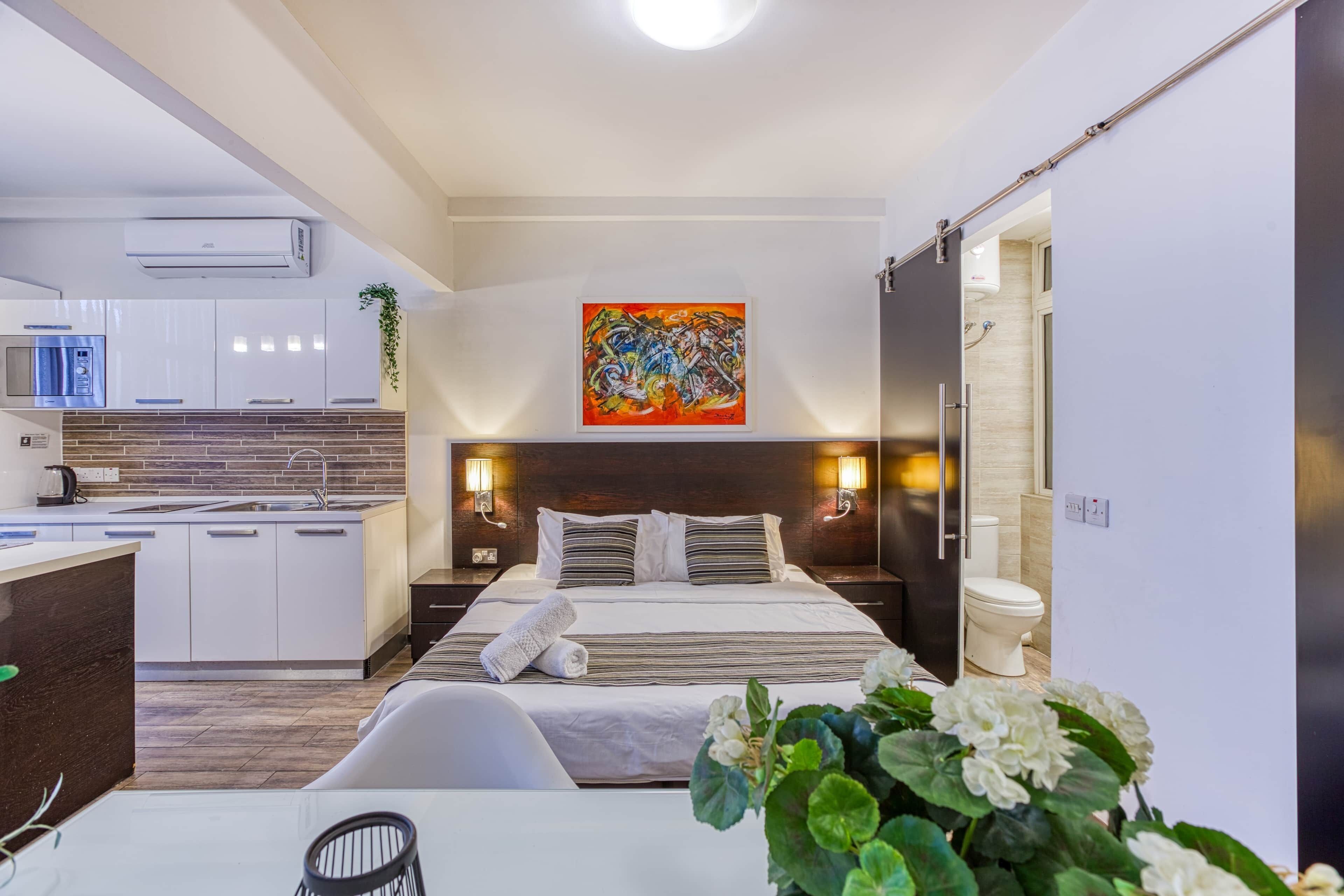 Une chambre d’hôtel moderne équipée d’une cuisine, d’un grand lit, de tables de chevet et d’un tableau au mur.