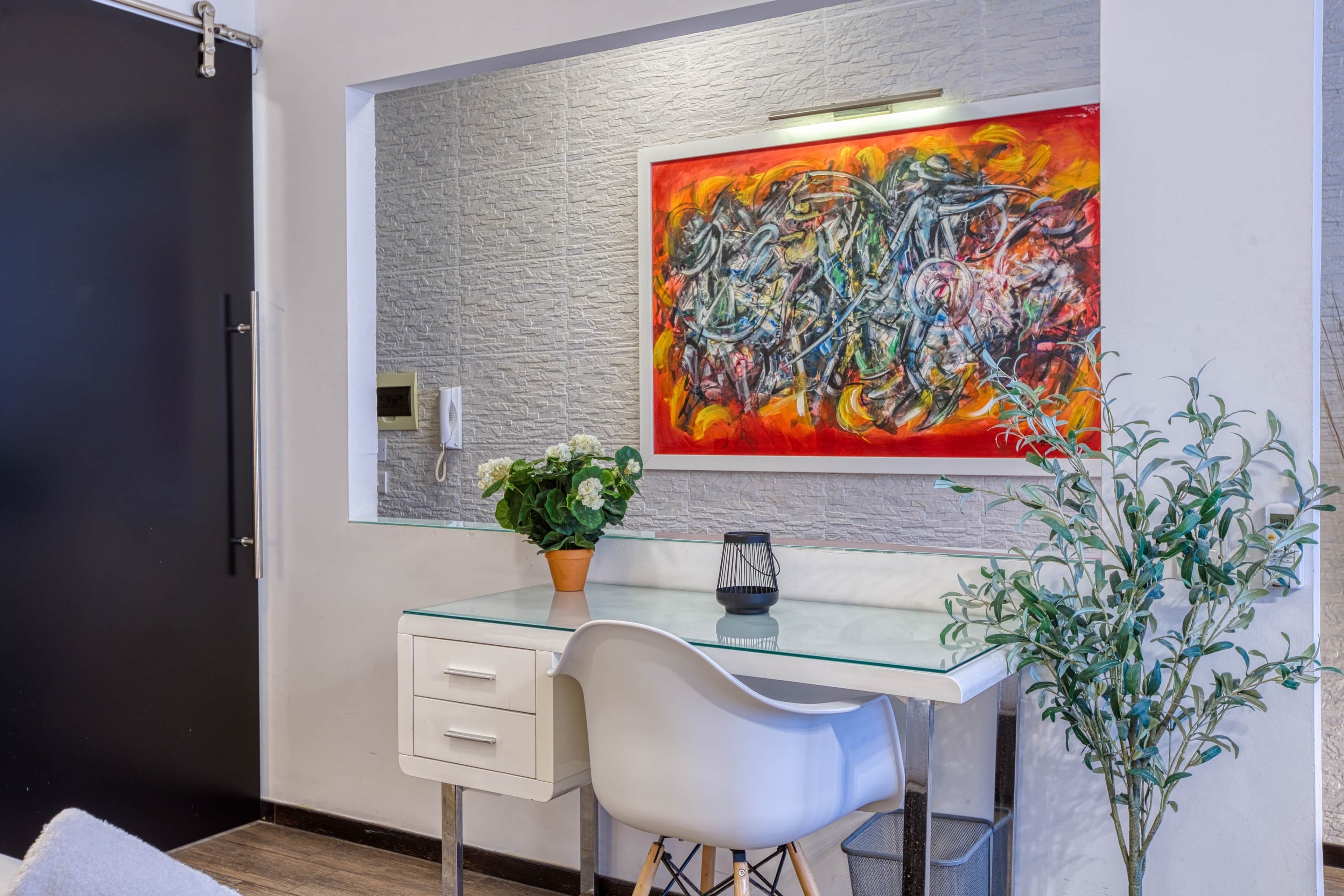 Quarto moderno com escrivaninha de vidro, cadeira branca, planta em vaso e uma grande pintura abstrata na parede.