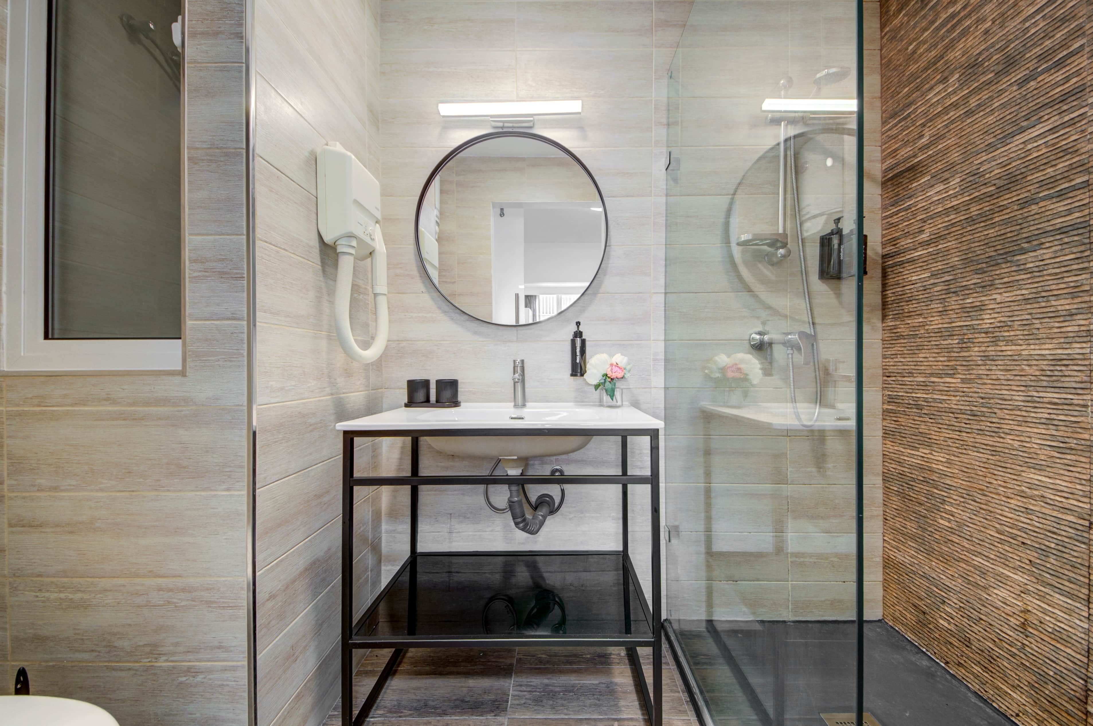 A modern bathroom with a glass shower enclosure, a round mirror, and a white sink with a black frame.