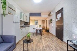 Eine kleine Küche mit weißen Schränken, einer Mikrowelle und einem Kühlschrank. Es gibt einen Essbereich mit Tisch und Stühlen, und ein Sofa im Vordergrund.