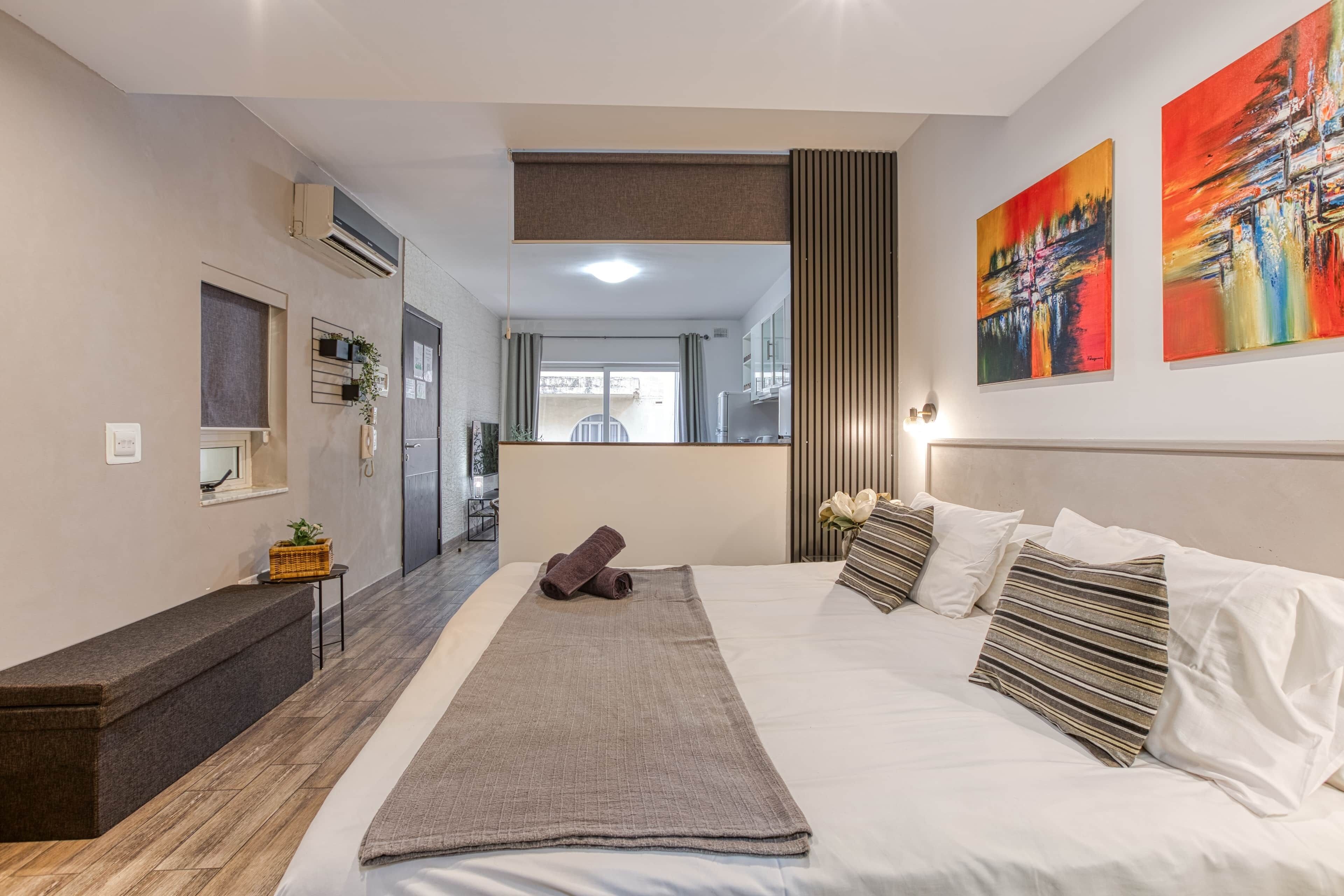 A modern hotel room with a large bed, a bench, a small table, and a view of the outside.
