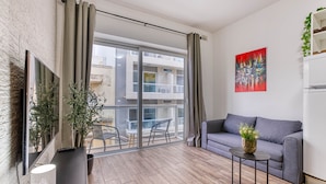 A modern living room with a grey sofa, a small side table, a TV mounted on a brick wall, and a sliding glass door leading to an outdoor balcony. - St. Julian's Studio 301 - Near Beach & Paceville (Saint Julian's)
