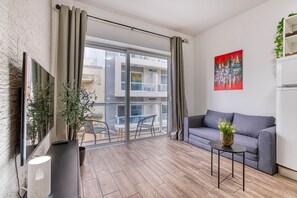 Ein modernes Wohnzimmer mit grauem Sofa, einem kleinen Beistelltisch, einem an einer Ziegelwand montierten Fernseher und einer Glastür, die auf einen Außenbalkon führt.