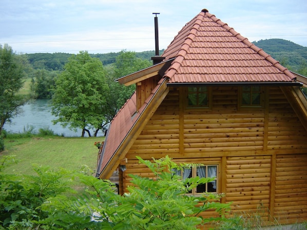 A wooden cabin with a sloped roof, a window, and a view of a river and forest.