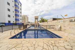 A swimming pool with stone tiles, a metal railing, and a view of a multi-story building with balconies. - Apto com Piscina e Vaga a 750m da Praia da Enseada (Bertioga)
