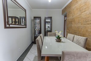 A dining area with a white table, cushioned chairs, a mirror, and display cabinets. - Apto com Piscina e Vaga a 750m da Praia da Enseada (Bertioga)
