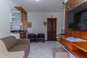 A living room with a sofa, two chairs, a TV, a wooden cabinet, and a kitchen area with a sink and cabinets. - Apto com Piscina e Vaga a 750m da Praia da Enseada (Bertioga)