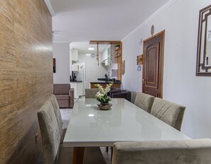 A dining area with a long table, chairs, a vase with flowers, a kitchen in the background, and a wooden door. - Apto com Piscina e Vaga a 750m da Praia da Enseada (Bertioga)