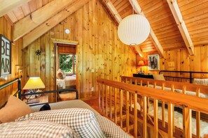 En stuga med träpanel på insidan, innehållande en säng, en lampa, en inramad tavla och en trappa.