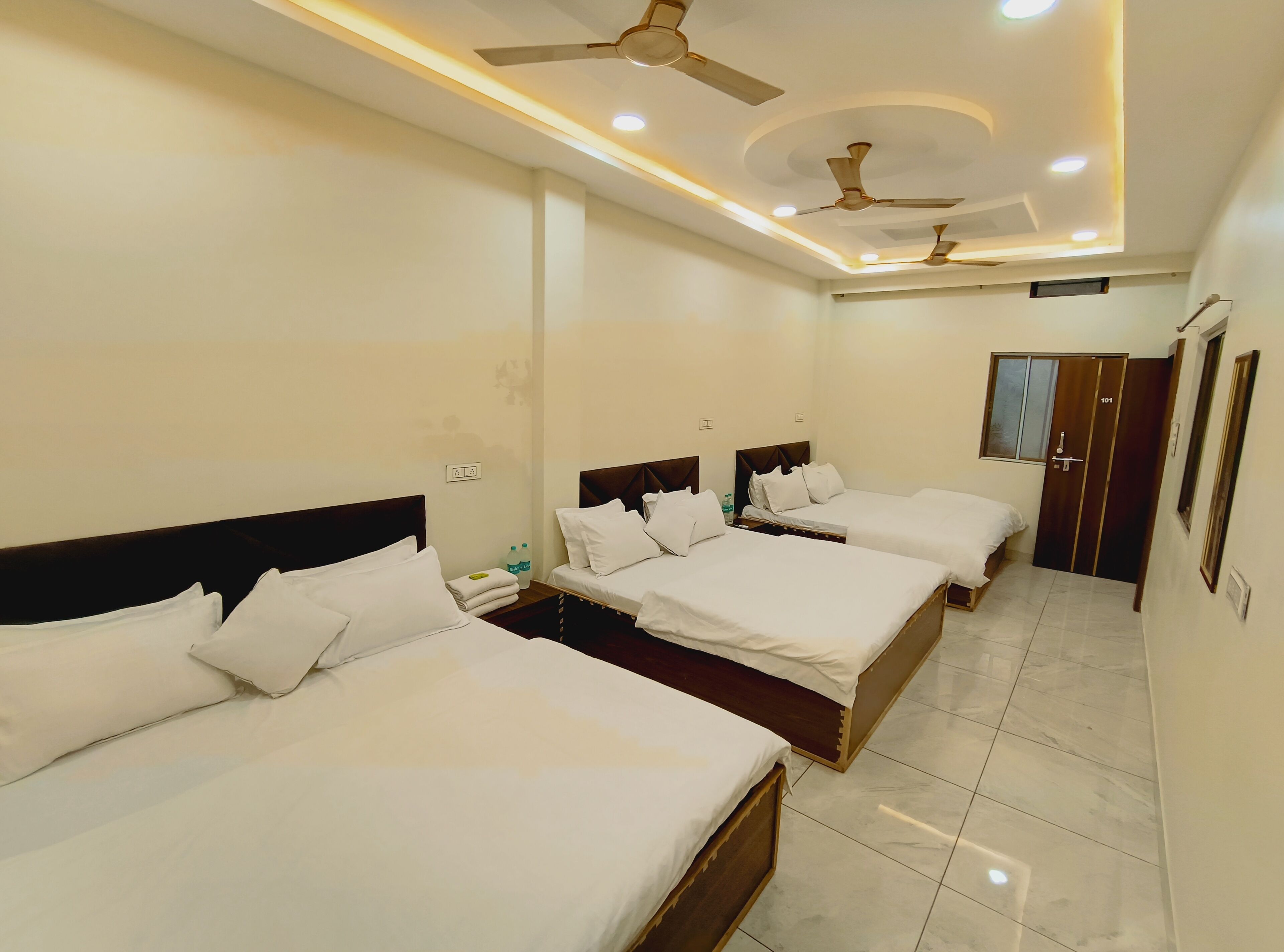 A hotel room with three beds, ceiling fans, recessed lighting, a water dispenser, and a door with a number.