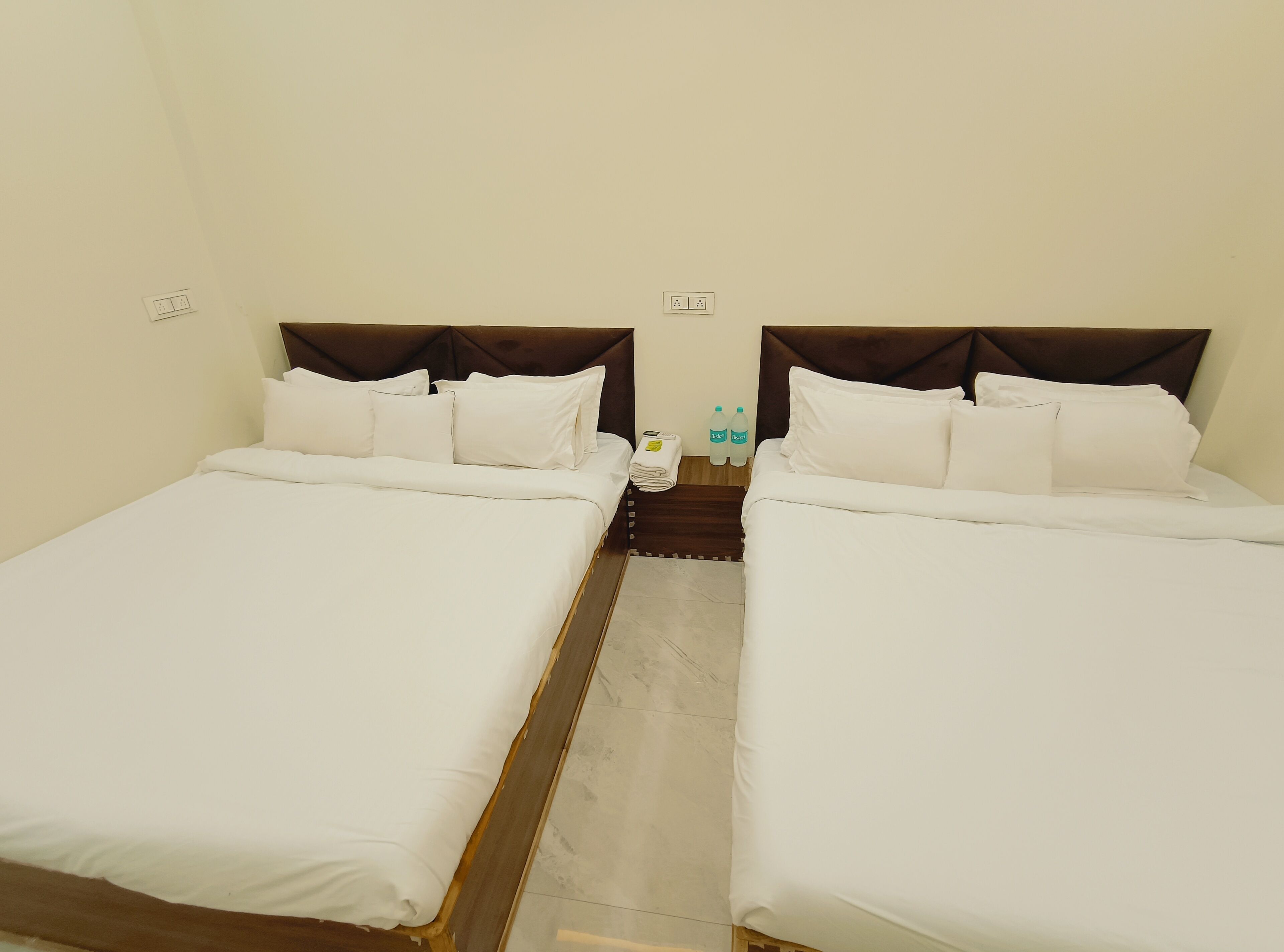 Hotellihuone, jossa on kaksi erillistä sänkyä, puinen pääty, pieni pöytä, jossa on pullo ja pyyhe, sekä pelkistetty seinä, jossa on pistorasia.