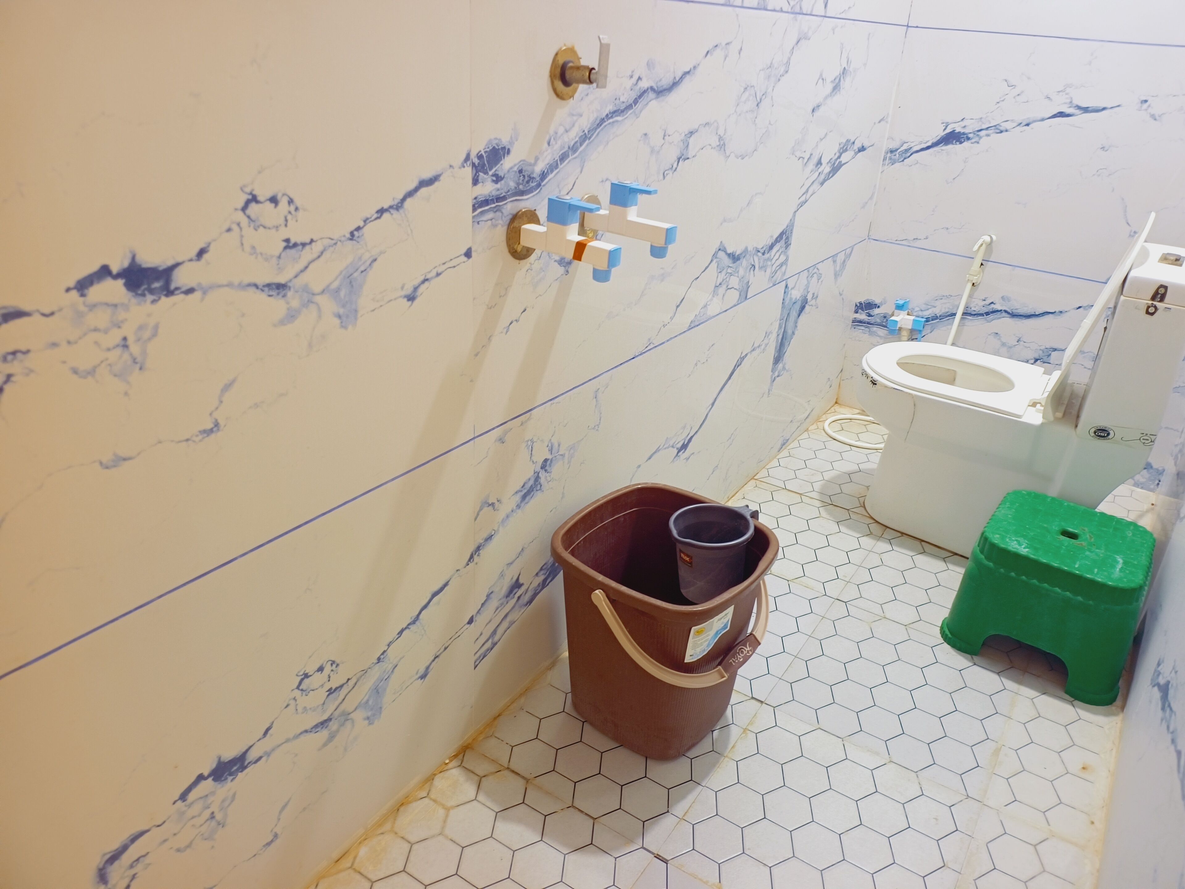 A bathroom with a toilet, a bucket, and a green stool.