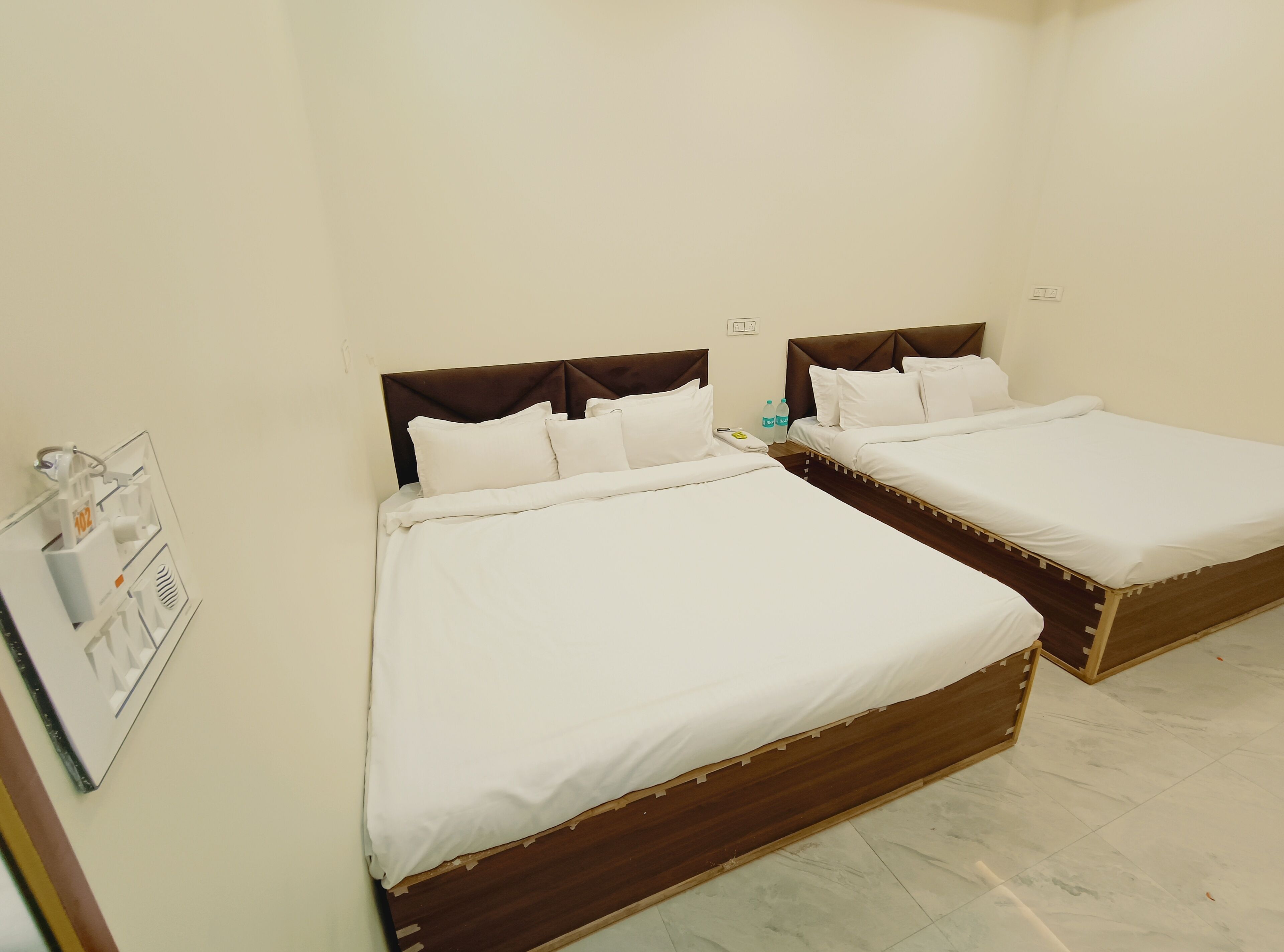 Ein Hotelzimmer mit zwei Betten, einem Nachttisch, einem an der Wand montierten Lichtschalter und einem kleinen Regal mit Toilettenartikeln.
