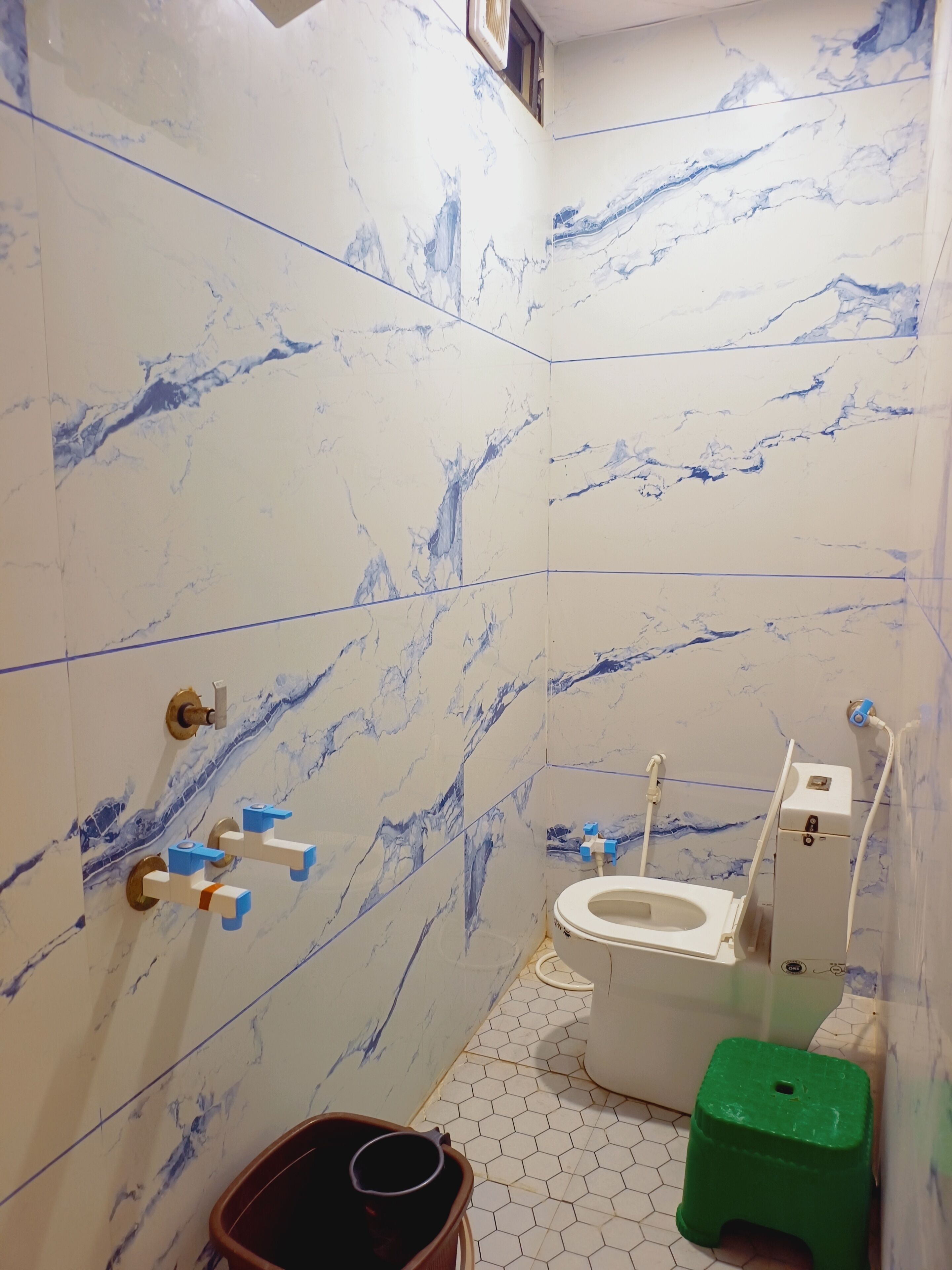 A bathroom with marble walls, a toilet, a green stool, and a bucket.