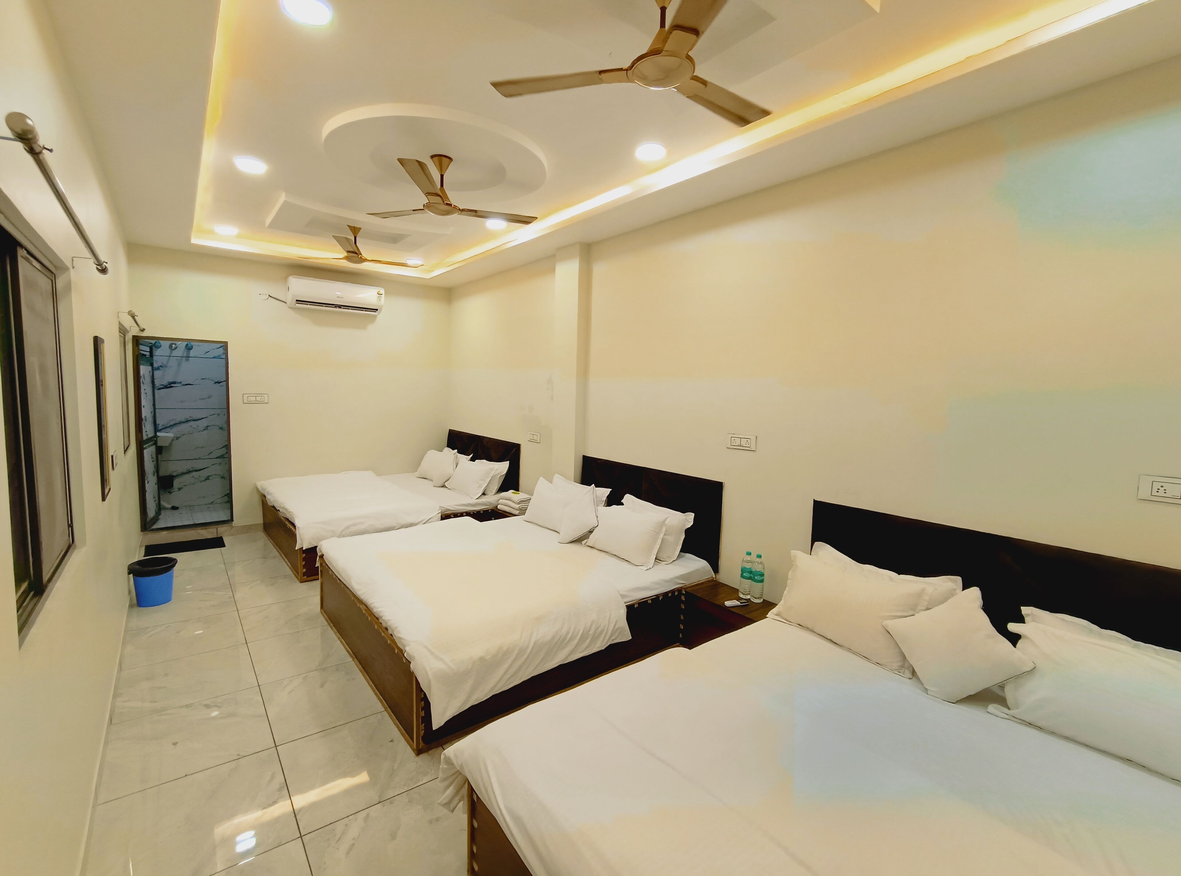 Habitación de hotel con tres camas, ventiladores de techo, luces empotradas, baño con ducha de mármol y un cesto de basura azul.