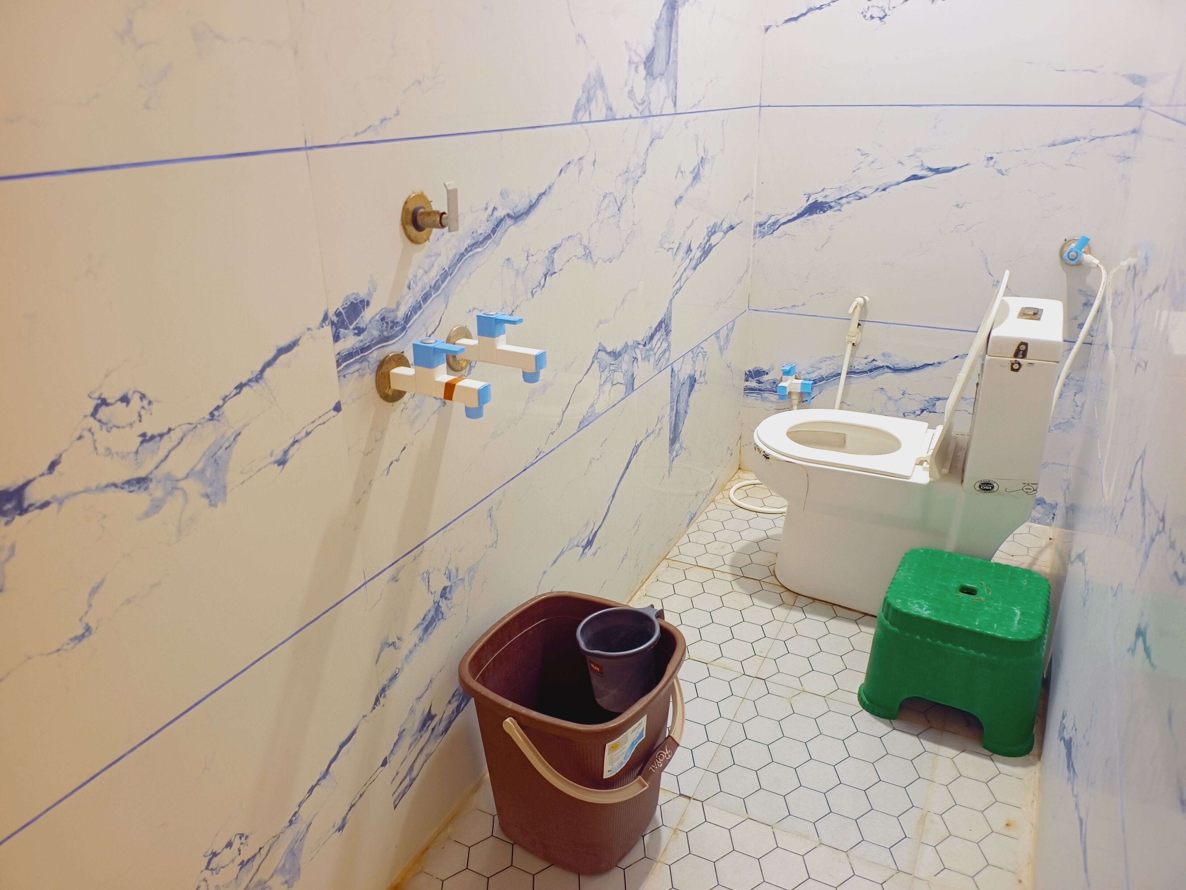 A bathroom with marble walls, a toilet, a green stool, and a brown bucket.