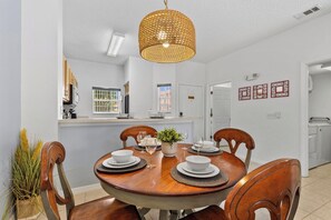 A dining room with a round table set for four, a hanging light fixture, and a kitchen in the background. - Sleeps 6·BBQ·Game Table·Clubhouse Amenities (Kissimmee)