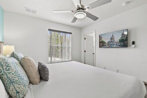 Ein Schlafzimmer mit einem großen Bett, einem Deckenventilator, einem Fenster mit Jalousien, einer Tür und einem an der Wand montierten Fernseher.