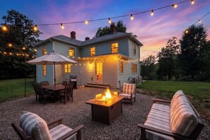 Une maison à deux étages dotée d’un patio aménagé avec des meubles d’extérieur, un foyer et des guirlandes lumineuses.