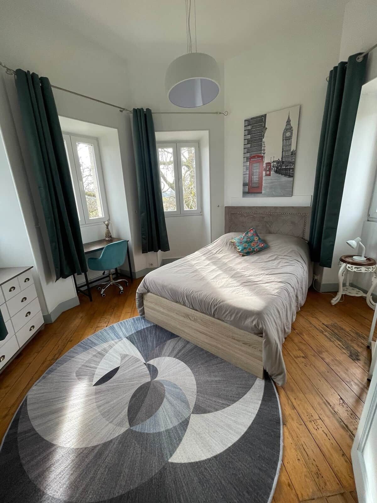 Een slaapkamer met een bed, een bureau, een stoel, een lamp en een schilderij van een stadsgezicht.
