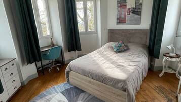 Een slaapkamer met een bed, een bureau, een stoel, een lamp en een schilderij van een stadsgezicht.