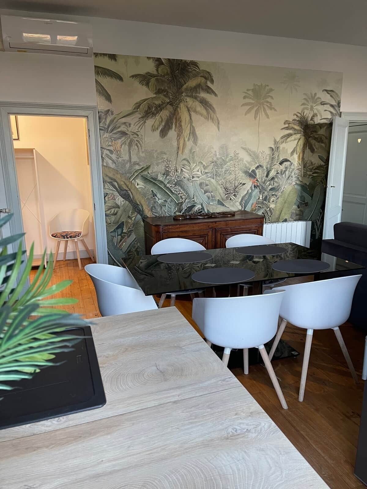 Uma área de refeições com um mural tropical, uma mesa de madeira, cadeiras brancas e uma pequena mesa lateral.
