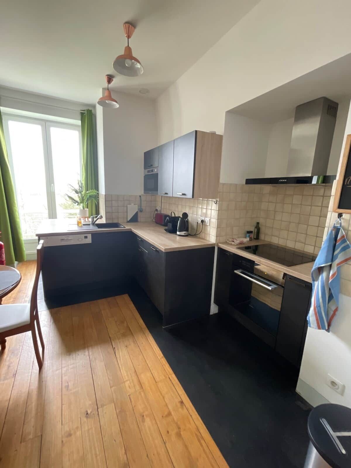 Una cocina compacta con suelo de madera, una mesa de comedor, horno empotrado y una ventana con cortinas.