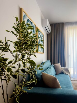 Ein modernes Wohnzimmer mit einem türkisfarbenen Sofa, einer großen Pflanze und gerahmten Kunstwerken an der Wand.
