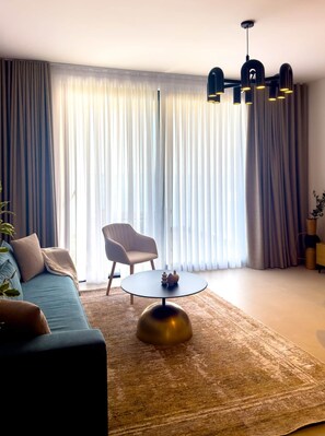 Ein modernes Wohnzimmer mit einer Couch, einem runden Couchtisch, einem Sessel und einem großen Fenster mit Vorhängen.