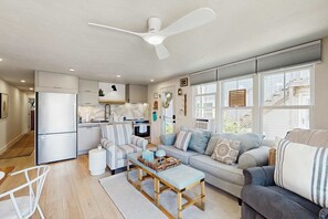 Una cocina moderna con refrigerador empotrado, una zona de comedor con mesa y sillas blancas, un rincón de descanso con sofá y reposapiés, y ventanas grandes con persianas.