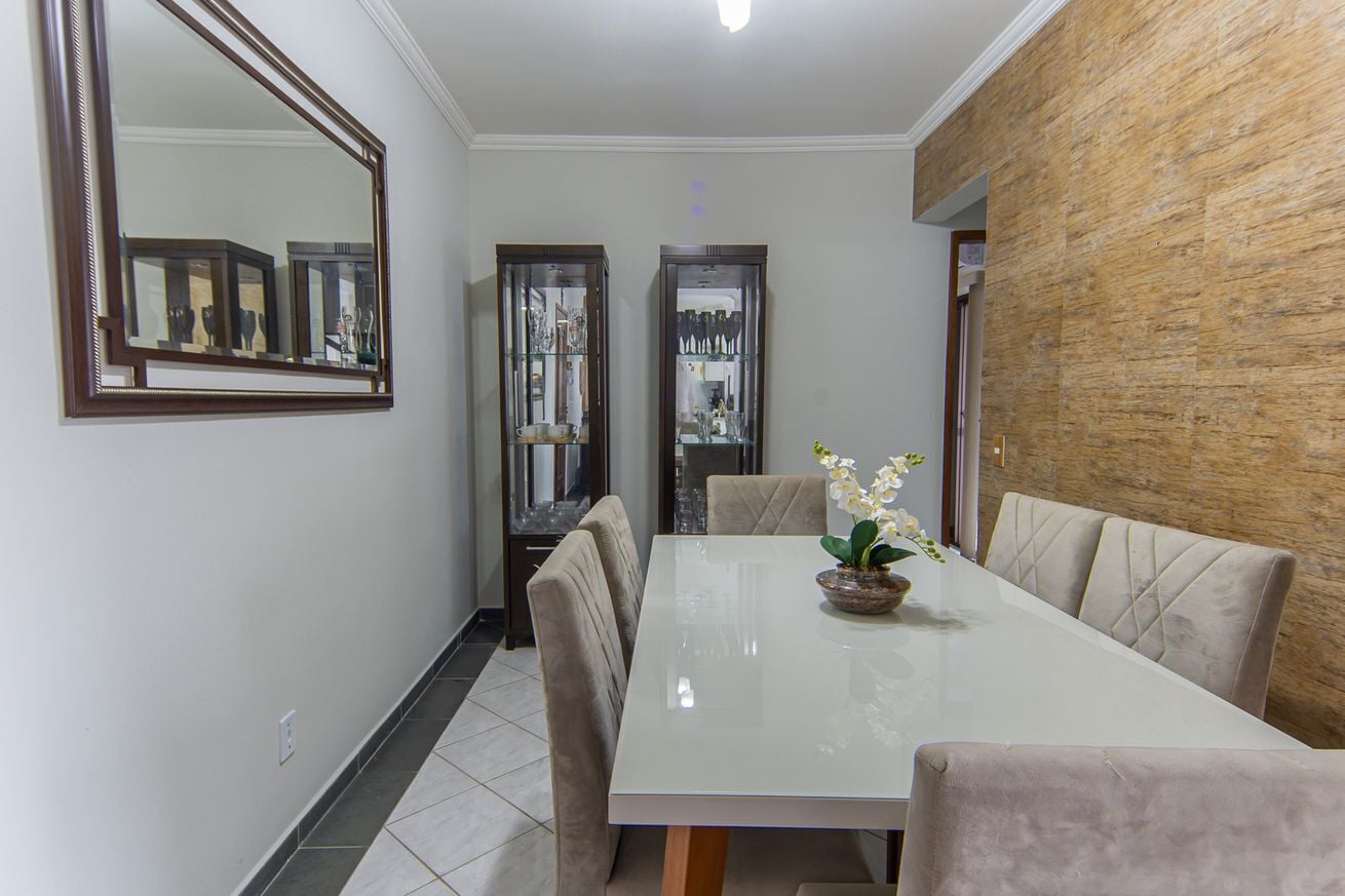 Uma sala de jantar com uma mesa branca, cadeiras acolchoadas, um espelho e armários de exposição.