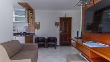 Una sala de estar con un sofá, dos sillones, un televisor, un mueble de madera y una zona de cocina con un lavabo y armarios.