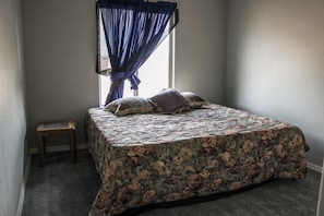 Quarto com cama, um banquinho e janela com cortinas azuis.