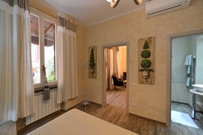 A hotel room with a wooden floor, a large window with curtains, a radiator, and a painting of potted plants on the wall. - San Francesco Ripa 2 - Fornarina Oasis Cottage (Rome)