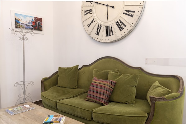 Un canapé vert agrémenté d’un coussin à motifs, une horloge murale avec des chiffres romains et un lampadaire.