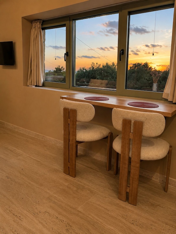 Una habitación con una mesa de madera y dos sillas, un televisor montado en la pared y una ventana con vista al atardecer.