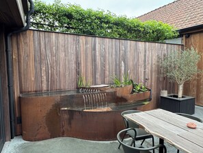 Une clôture en bois, un élément décoratif avec de l’eau, une table avec des chaises et une plante en pot.