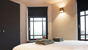 Une chambre à coucher avec un grand lit, une fenêtre avec des stores, une lampe fixée au mur et une porte.