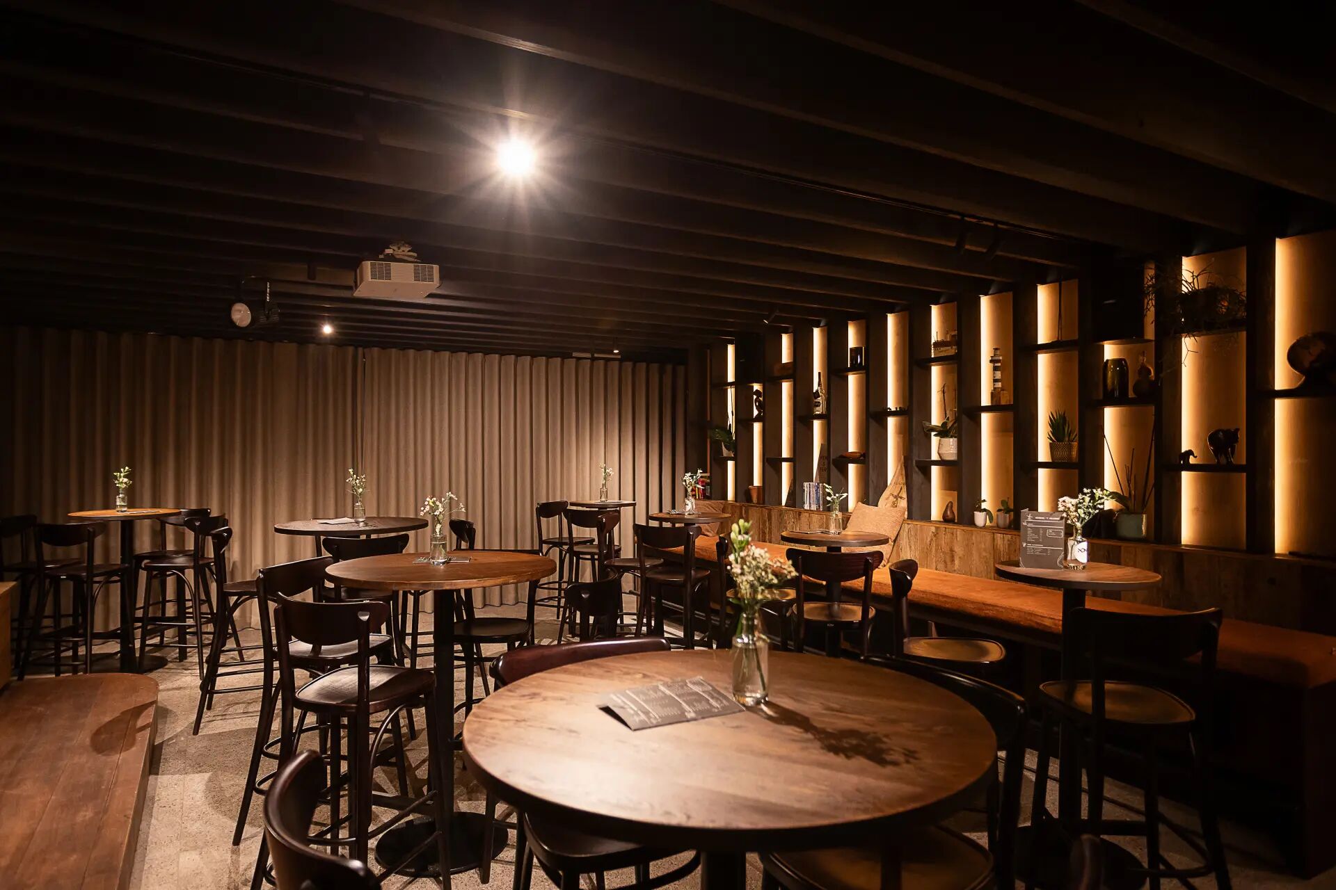 A dimly lit restaurant with round tables and chairs, wooden beams on the ceiling, and shelves with various items on the walls.