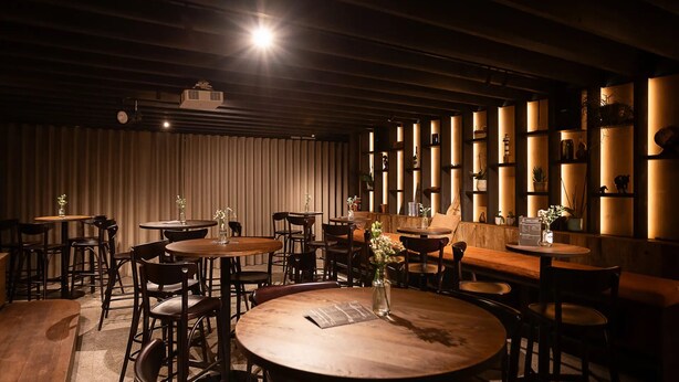 Un restaurant à la lumière tamisée, avec des tables et des chaises rondes, des poutres en bois au plafond et des étagères ornées de divers objets le long des murs.