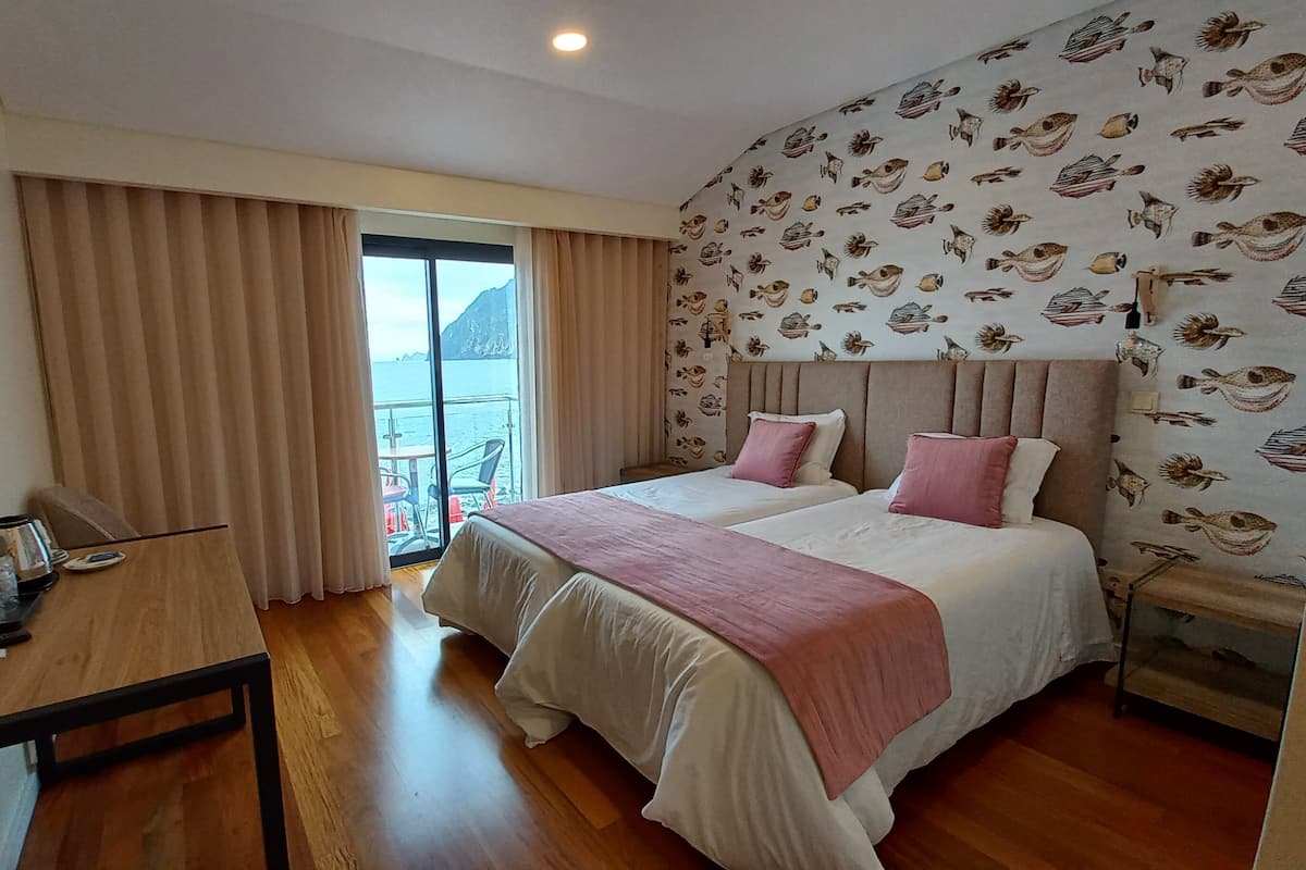 Een hotelkamer met twee bedden, een houten bureau, een televisie en een balkon met uitzicht op zee.