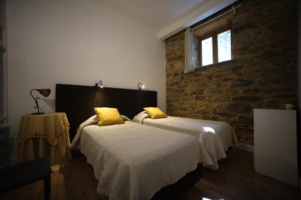 A bedroom with two beds, a stone wall, a window, and a table with a lamp.